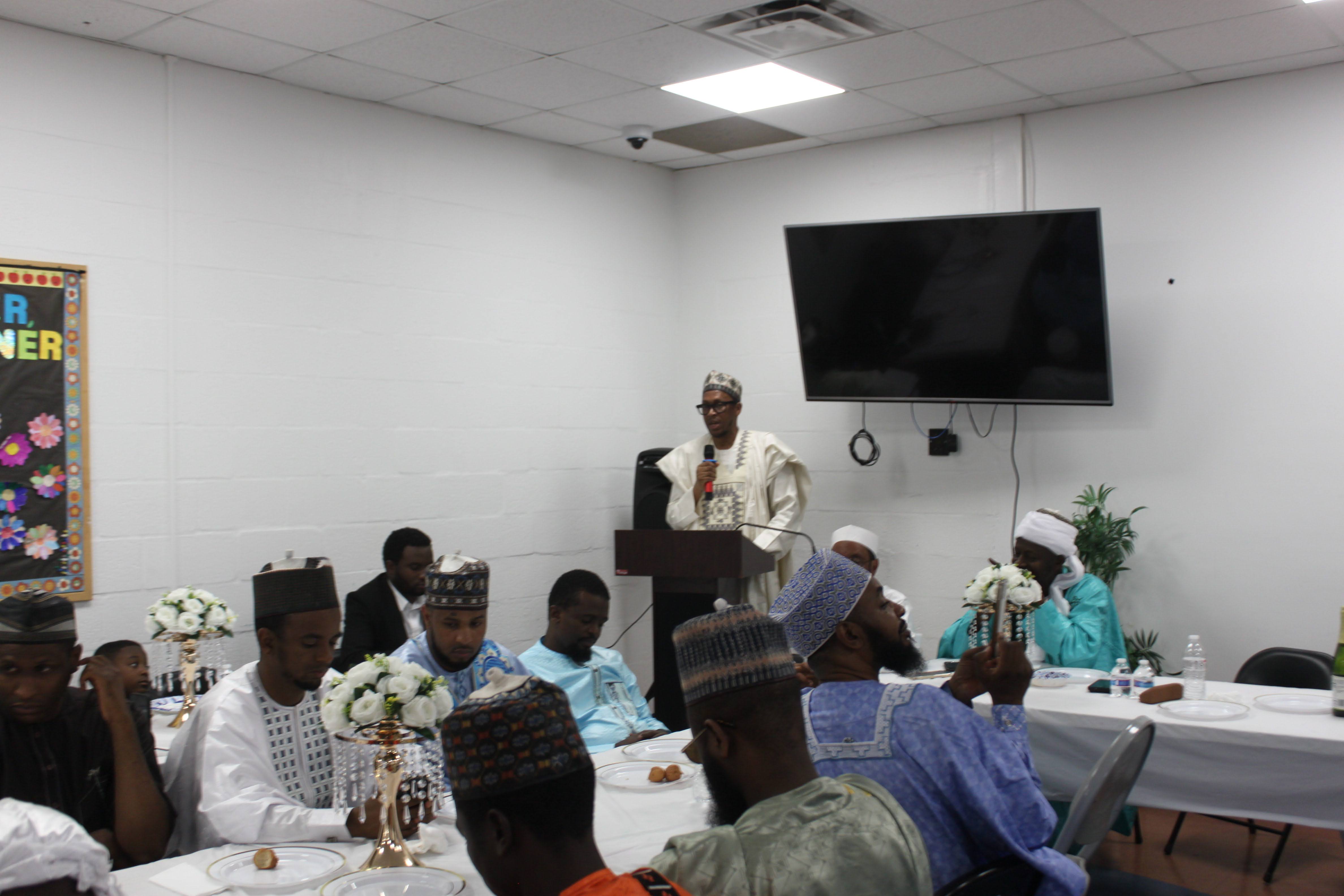 Wide view of the Al Ada community dinner hall with decorated tables and a speaker addressing seated guests in traditional dress