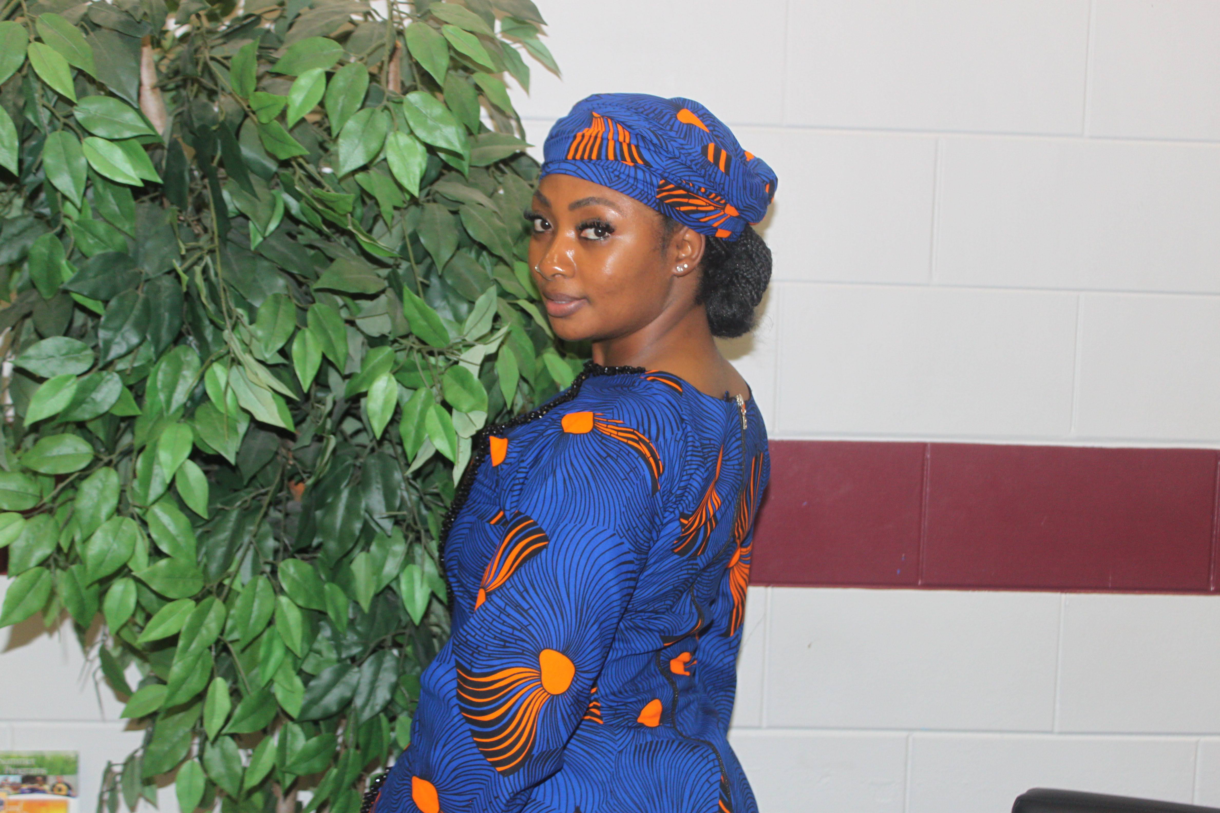 Fulani woman in a blue and orange dress and matching headwrap beside greenery